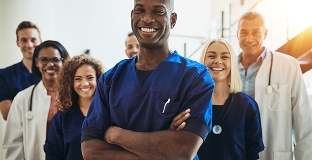 Young African male doctor smiling while standing in a hospital corridor with a diverse group of staff in the background