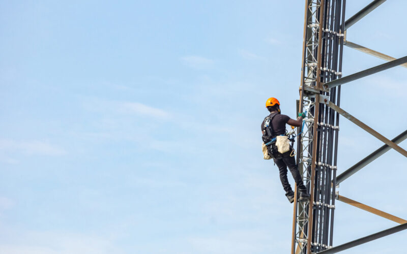 Communication maintenance. Technician climbing on telecom tower antenna against blue sky background, copy space.