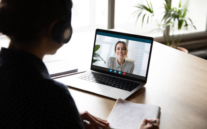 Back view of female employee sit at desk at home have Webcam conference on laptop with business partner or client, woman worker talk on video call with coworker or consultant, watch online webinar